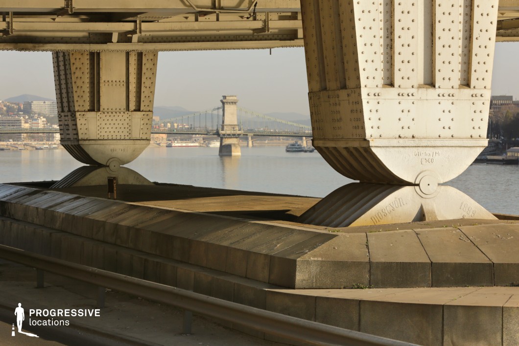 The image captures the robust iron pillars of a bridge, showcasing intricate riveted construction and a unique balancing mechanism at their base. The solid concrete foundation emphasizes the bridge's structural integrity, while the perspective draws the viewer’s eye through the frame toward another historic suspension bridge in the background. The soft lighting highlights the industrial textures of the metal and stone, adding depth to the composition. The water beneath reflects the urban landscape, enhancing the sense of stability and connection between old and modern engineering. This location is well-suited for film and photography requiring industrial aesthetics, architectural contrasts, or urban infrastructure themes.