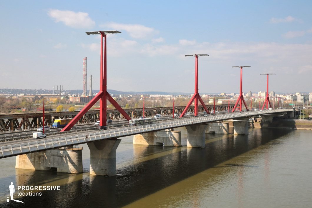 The image showcases a contemporary steel bridge with striking red support pylons spanning a wide river. The bridge's minimalist yet industrial design features clean geometric lines and large supporting piers that anchor the structure. The elevated perspective emphasizes the repetitive symmetry of the pylons, leading the viewer’s eye along the length of the bridge. In the background, an industrial landscape with factory chimneys and modern urban developments contrasts with the sleek design of the bridge. The river beneath reflects the bridge’s towering forms, adding depth to the scene. This location is ideal for filming sequences involving modern infrastructure, urban landscapes, or transport-related themes, providing a bold visual contrast between contemporary engineering and industrial history.