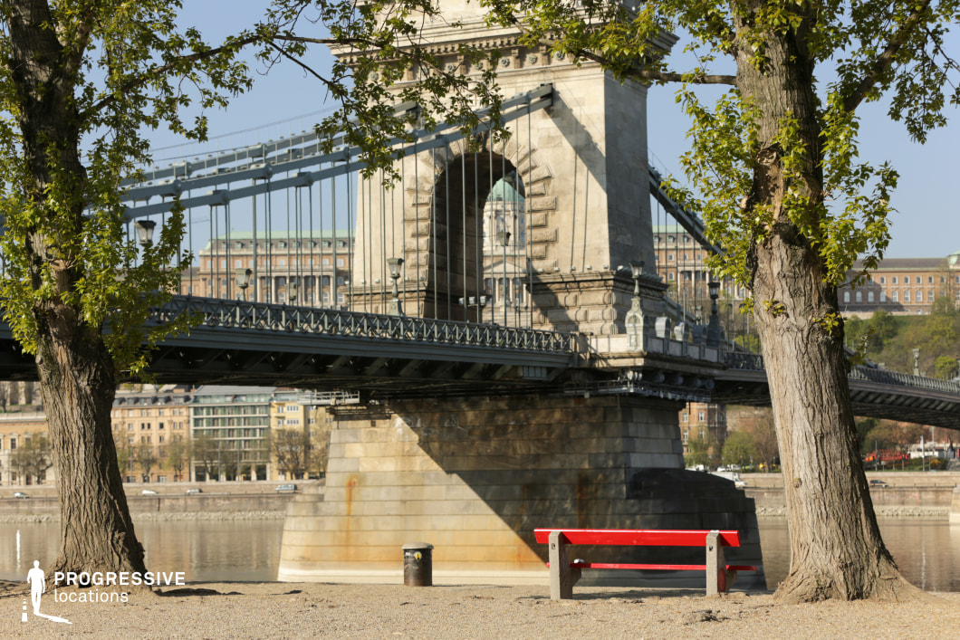 This photograph presents a grand suspension bridge with intricate stone pillars and steel cables, framed by two lush trees in the foreground. The bridge’s detailed architecture contrasts beautifully with the organic forms of the trees, creating a balanced composition. A red bench on a sandy riverbank adds a pop of color and provides a focal point in the lower section of the image. The bridge’s massive stone pier, visible in the center, showcases weathered textures with signs of rust and water exposure, emphasizing its historical and industrial character. In the background, classical buildings with ornate facades contribute to the urban atmosphere, adding a sense of timeless elegance. This location is perfect for capturing cinematic moments with a mix of natural and urban elements, offering a peaceful yet structured setting for storytelling or commercial shoots.