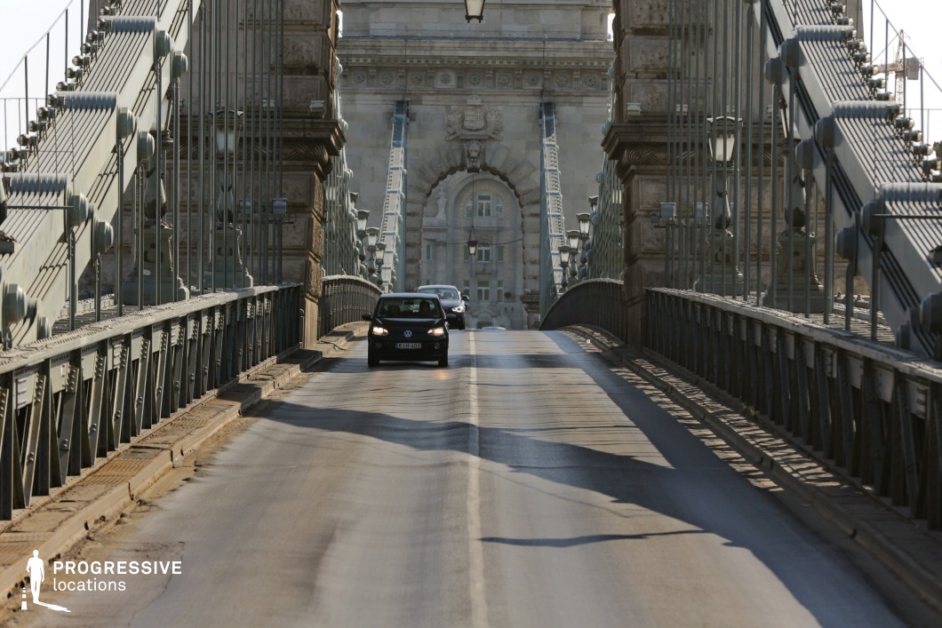 A grand suspension bridge with intricate steel cables and iron railings stretches across the frame, leading toward an imposing stone gateway adorned with detailed architectural elements. The linear perspective created by the bridge’s structure emphasizes depth, drawing the eye towards the central arch, which is embellished with classical stone reliefs. The bridge's road surface exhibits signs of wear, adding a layer of authenticity to the scene. The interplay of light and shadow enhances the texture of the iron framework, while the repeating pattern of suspension cables and street lamps contributes to the bridge's classical elegance. This setting is ideal for cinematic sequences requiring historical depth and urban grandeur, offering a visually compelling mix of engineering mastery and old-world architecture.