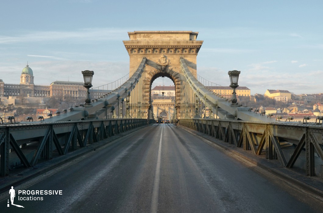 A grand suspension bridge with a symmetrical iron framework and stone gateway spans the scene, leading the viewer’s eye toward a central archway. The bridge’s metal structure, adorned with evenly spaced lampposts, contrasts with the solid stone architecture, creating a visually striking composition. Beyond the bridge, an expansive cityscape unfolds, featuring a historic palace with a domed roof, classical facades, and terraced buildings bathed in soft light. The alignment of the bridge with the distant structures adds depth and perspective, making it an ideal location for cinematic urban sequences or period productions. The bridge’s weathered iron framework and textured road surface add an authentic touch, emphasizing its historical significance. The combination of metal engineering and neoclassical architecture provides a balanced aesthetic, perfect for showcasing grandeur and urban heritage in a film or photography project.