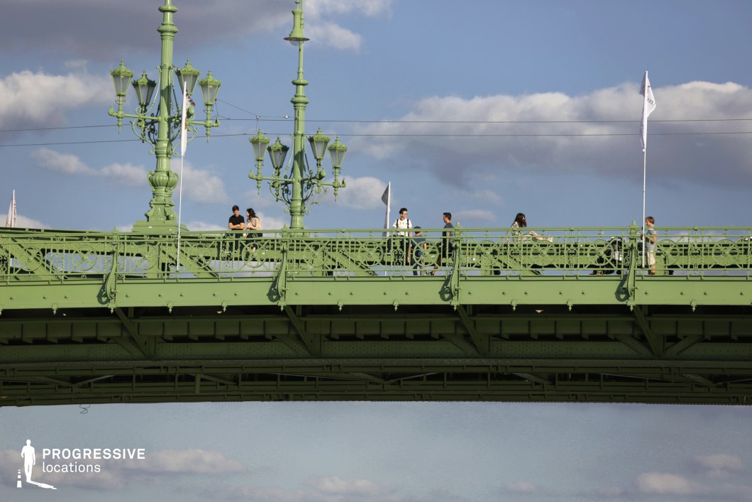 An intricately designed green metal bridge with elegant, vintage-style lampposts stands against a backdrop of a clear blue sky with scattered clouds. The structure showcases ornate railings with decorative ironwork, evoking a historical European aesthetic. Flags attached to the railing add a dynamic element, fluttering gently in the breeze. The underside of the bridge reveals its strong yet decorative framework, highlighting the detailed craftsmanship of the metal trusses. The combination of architectural detail and open sky makes this location an excellent choice for film and photography projects requiring a classic urban bridge setting with a touch of timeless elegance.