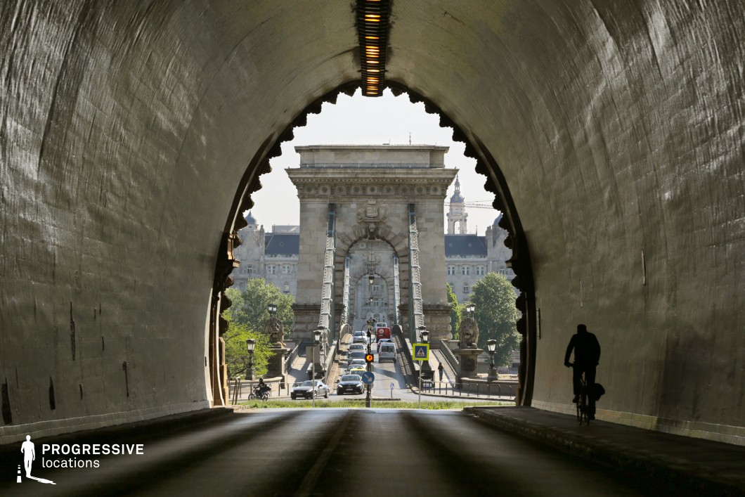 A grand stone tunnel with curved walls and a rugged, textured surface frames a historic suspension bridge in the distance. The tunnel's interior is dimly lit by a central row of small lights, creating a contrast with the bright daylight outside. Through the tunnel's arched opening, an ornate suspension bridge with massive stone pillars and intricate iron cables dominates the composition. Decorative elements such as lion statues, wrought-iron street lamps, and classical facades enhance the scene's architectural richness. This location offers a cinematic contrast between light and shadow, perfect for dramatic urban storytelling or historical-themed productions.