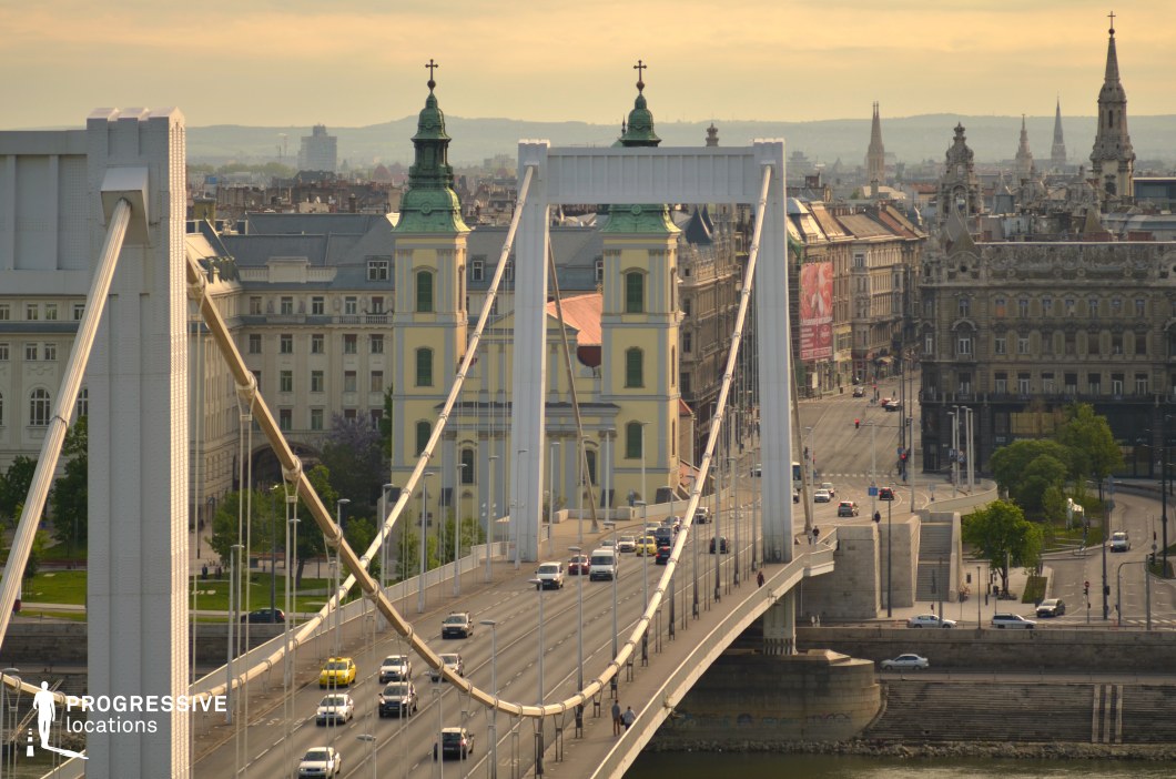 A striking composition of a modern white suspension bridge leading into a historic cityscape, creating a fusion of architectural styles. The bridge’s towering white pillars and taut cables form elegant curves, contrasting with the ornate facades of classical buildings in the background. A soft golden light bathes the scene, emphasizing intricate details of the historic structures, including a prominent yellow twin-towered church with green domes. The bridge’s roadway, lined with evenly spaced lampposts, adds depth and perspective, drawing the eye toward the city's urban core. The surrounding streets are framed by grand, historic buildings with intricate facades, spires, and decorative elements, reinforcing the juxtaposition of contemporary infrastructure and classical elegance. The image captures an ideal balance of modern engineering and old-world charm, making it a visually compelling scene for urban-themed creative projects.