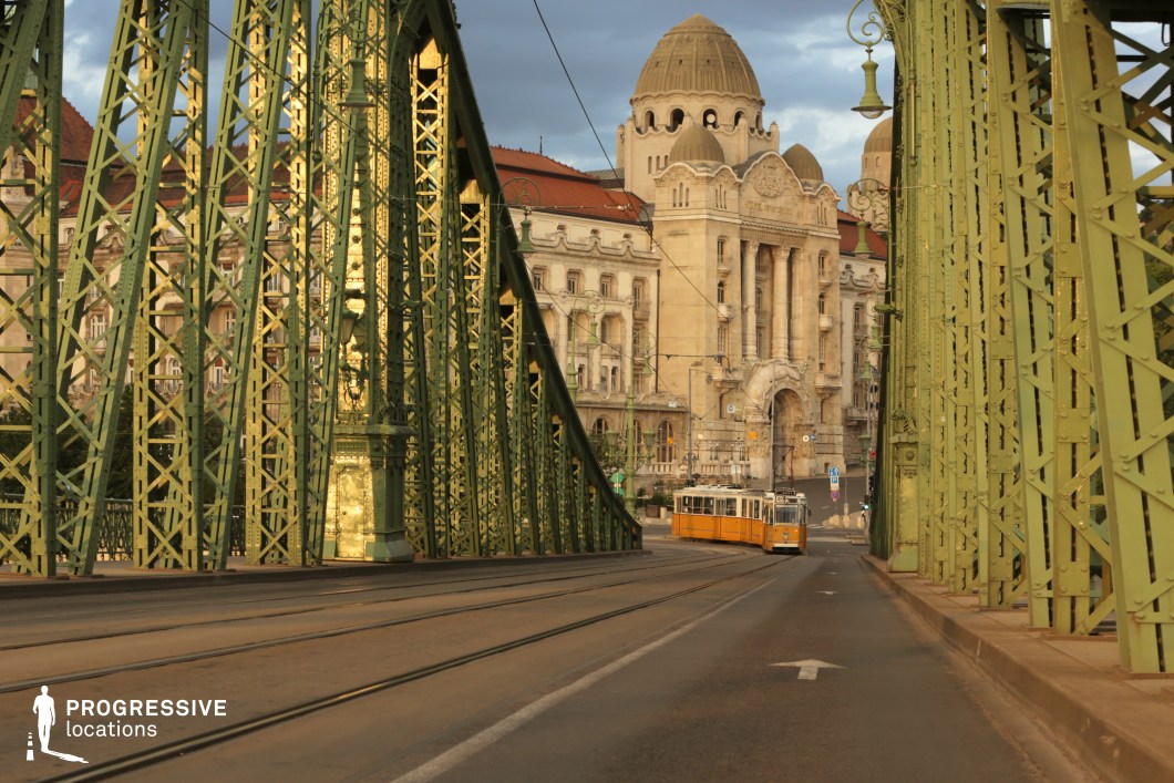 An ornate green iron bridge with intricate latticework spans across an urban setting, leading towards a grand historic building with domed towers and an elaborate façade. The bridge’s detailed metal framework, painted in a deep green hue, contrasts with the golden sunlight reflecting off its surfaces. Tram tracks run along the road, guiding a classic yellow tram that adds a vibrant pop of color. Overhead lamps and electric cables contribute to the vintage atmosphere, while the background architecture emphasizes a mix of historical and Art Nouveau influences. This location is ideal for transportation-themed visuals, urban storytelling, and classic European cityscapes.