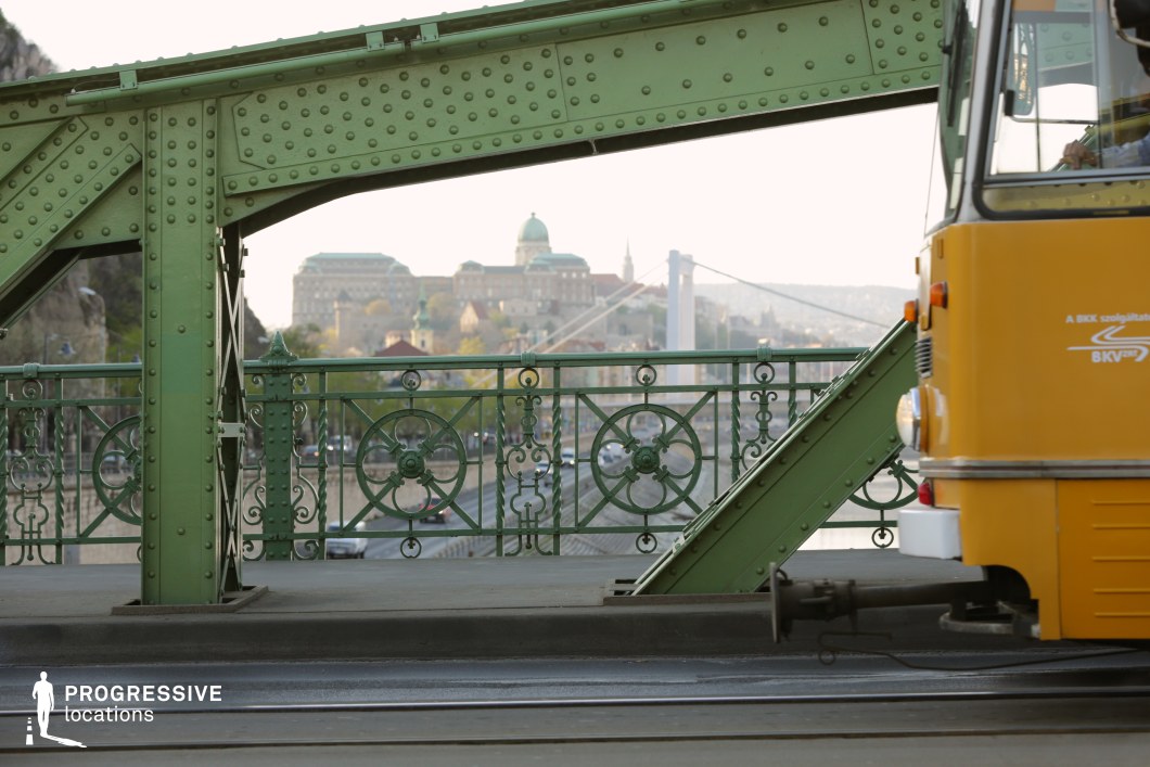A partially visible yellow tram passes over an ornate green iron bridge, showcasing intricate riveted metal beams and decorative railing details. The composition frames a scenic city backdrop with a domed historic building and a white suspension bridge in the distance. The tram, with its classic design, adds a dynamic urban element to the structure’s vintage aesthetic. The contrast between the warm tones of the vehicle and the cool green of the ironwork enhances the visual depth, making this setting ideal for showcasing historic European transport and architectural elegance.