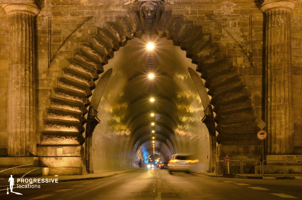 A monumental stone tunnel entrance framed by an intricately detailed arch with bold geometric patterns and a sculpted lion's head keystone. The tunnel extends into the distance, with a series of warm, evenly spaced overhead lights creating a rhythmic visual effect. The smooth, curved interior walls contrast with the ornate and textured stone façade, which features classical columns and decorative stonework. The tunnel’s perspective draws the eye into the depth of the scene, making it an ideal setting for dramatic urban sequences, cinematic chase scenes, or historical architectural studies.