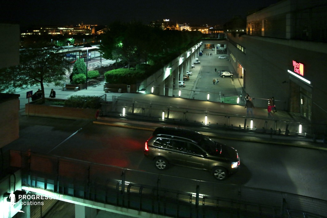 A modern, multi-level urban space illuminated by artificial lighting, featuring an elevated roadway, pedestrian walkways, and landscaped public areas. The geometric design of the architecture is complemented by linear lighting along the railings, creating a structured, contemporary aesthetic. The scene is framed by a commercial building with a neon-lit entrance, while in the background, a cityscape with historic elements adds depth and contrast. The layered composition and various elevation changes make this an ideal location for urban storytelling, architectural photography, or night-time cinematic sequences.