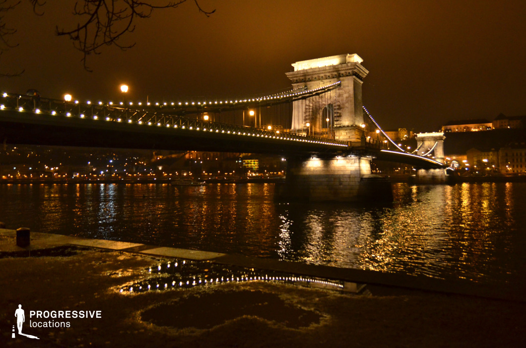 A grand suspension bridge stretches across a calm river, adorned with glowing string lights that reflect on the water’s surface. The massive stone pillars and intricate ironwork of the bridge are highlighted by warm architectural lighting, emphasizing its historical significance. The background features a softly lit cityscape, adding depth to the composition. The riverside foreground introduces texture, with reflections creating a visually dynamic contrast between the built environment and natural elements. This setting is ideal for cinematic night scenes, architectural photography, or projects requiring an elegant and atmospheric urban riverfront backdrop.