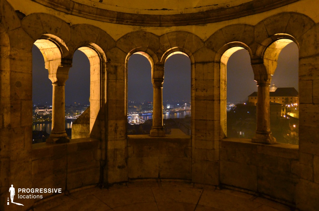 A stunning nighttime cityscape viewed through a series of aged stone arches, creating a visually rich and historic perspective. The elegant, symmetrical arches feature classical architectural details, including rounded columns and weathered stone surfaces, enhancing the timeless atmosphere. Beyond the arches, the illuminated city skyline and a river with bridges add depth and contrast, while the interplay of warm interior lighting and cool night tones accentuates the scene’s dramatic composition. This location is ideal for cinematic framing, historical documentaries, or projects requiring a unique architectural vantage point over a vibrant urban landscape.