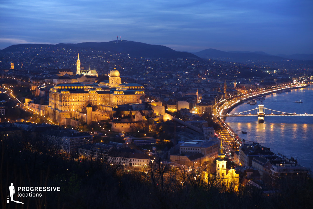 A breathtaking panoramic view of a historic city illuminated by warm golden lights, showcasing a grand palace, a gothic-style church, and a gracefully curving riverside boulevard. The suspension bridge, adorned with string lights, spans the river, connecting the urban landscape. The city's layout, with its blend of classical and baroque architecture, contrasts beautifully against the rolling hills in the background. The elevated perspective emphasizes the depth and grandeur of the city, making this an ideal setting for cinematic aerial shots, travel documentaries, or projects requiring a striking, atmospheric urban vista.