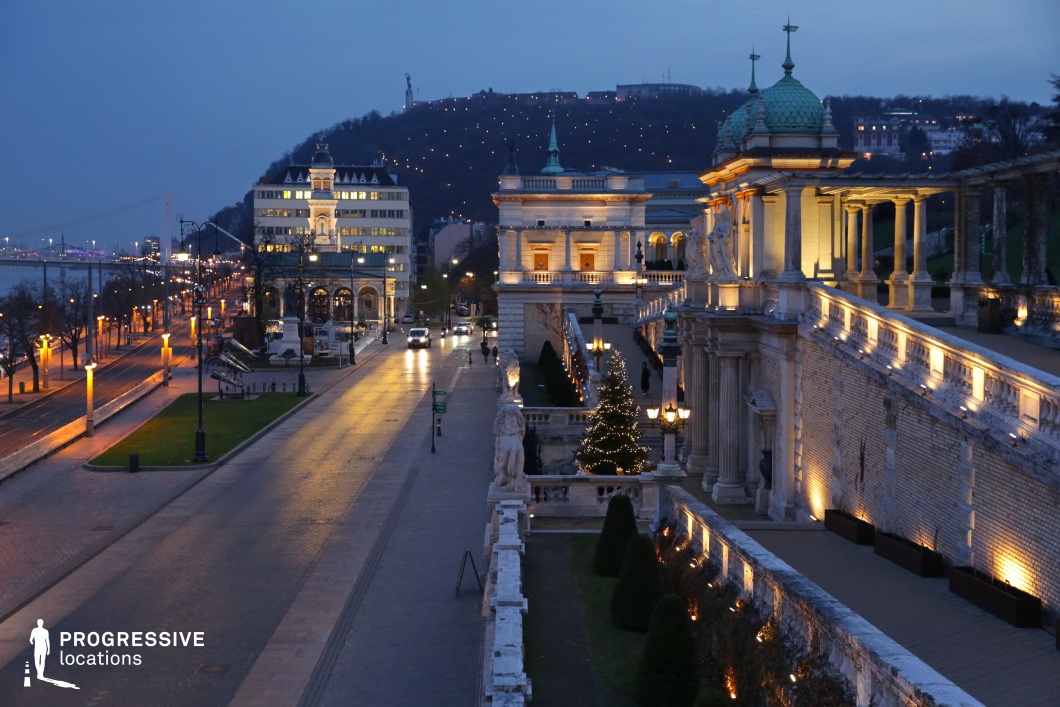 A refined urban promenade lined with grand neoclassical architecture, intricate balustrades, and ornate sculptures. The scene is bathed in soft, warm lighting, emphasizing the intricate details of the historic structures. Elegant street lamps and decorative statues contribute to the regal ambiance, while the road leads towards a distant bridge and a hillside dotted with lights, adding depth to the composition. The combination of historic and modern elements makes this an ideal location for period film settings, upscale urban photography, or projects requiring a sophisticated and atmospheric European cityscape.