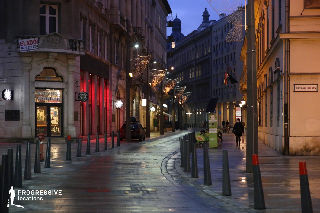 A stylish European pedestrian street lined with historic buildings featuring intricate facades, arched entrances, and decorative cornices. The pathway is paved with smooth cobblestones, bordered by evenly spaced bollards that guide the flow of movement. Elegant streetlights and festive decorative lighting create a warm ambiance, complementing the soft evening illumination. The street curves gently, leading towards a distant mix of classical and modern buildings, adding depth to the composition. This location is ideal for urban lifestyle photography, upscale commercial shoots, or film projects requiring a sophisticated and atmospheric city backdrop.