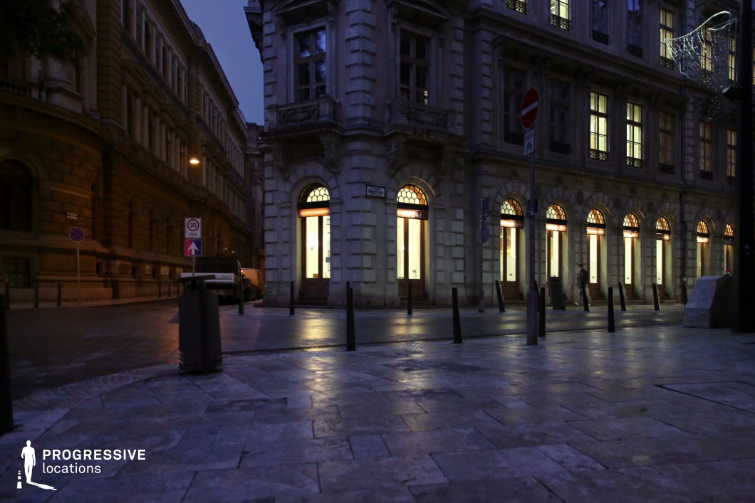 A striking neoclassical corner building with grand arched windows and intricate stone detailing, illuminated from within. The well-preserved façade features decorative cornices and sculpted accents, reflecting a timeless European elegance. The surrounding urban square is paved with large stone tiles, leading towards adjacent historic structures that enhance the depth of the scene. The ambient lighting and symmetrical composition create a refined and cinematic atmosphere, making this location ideal for period film productions, upscale urban photography, or projects requiring a sophisticated and architecturally rich backdrop.