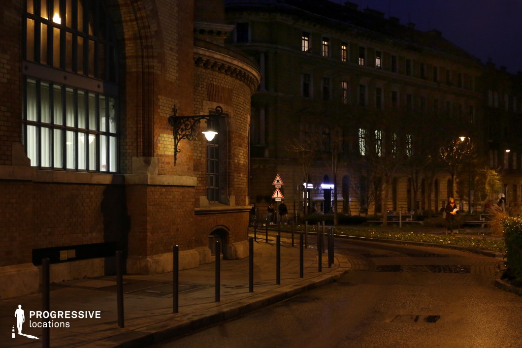 A dimly lit, curving street framed by historic brick buildings with arched windows and intricate wrought-iron lanterns. The soft glow from the streetlights contrasts with the dark tones of the architecture, creating a moody and cinematic atmosphere. The paved walkway, lined with short metal bollards, guides the viewer’s eye towards the background, where more historic buildings and a small park area add depth to the scene. The mix of old-world craftsmanship and urban night lighting makes this an ideal setting for noir-style cinematography, period dramas, or projects capturing the essence of a historic European city at night.