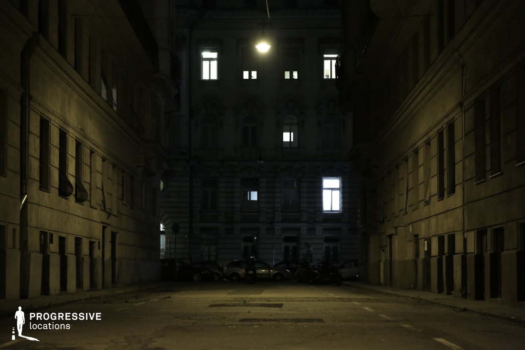 A dimly lit, enclosed alleyway flanked by aged buildings with worn facades and barred windows, creating a tense and mysterious atmosphere. A single overhead streetlight casts a faint glow, barely illuminating the cracked pavement and distant parked cars. The windows of the surrounding buildings reveal sparse interior lighting, adding to the sense of isolation. The composition, with its deep shadows and confined space, makes this an ideal setting for psychological thrillers, neo-noir cinematography, or urban horror storytelling.