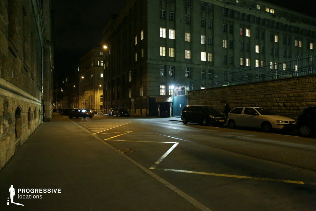 A dimly lit urban street lined with austere, institutional buildings featuring rigid geometric facades, tall barred windows, and cold concrete surfaces. The atmosphere is heavy with a sense of surveillance, reinforced by the imposing structures and a high metal fence enclosing one of the buildings. Sparse streetlights cast a harsh glow on the pavement, emphasizing the stark contrast between illuminated windows and the deep shadows of the street. The muted color palette and the absence of vibrant life make this an ideal setting for dystopian narratives, Cold War-era thrillers, or projects evoking themes of isolation and control.