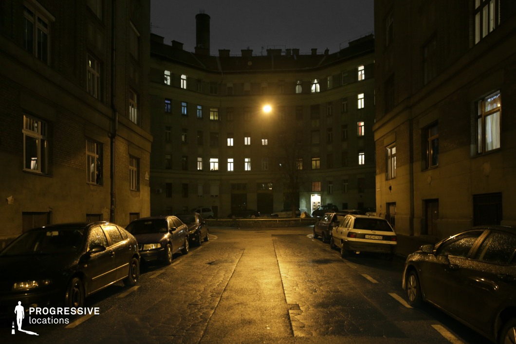 A dimly lit, enclosed courtyard surrounded by curved, utilitarian apartment buildings with uniform facades, small windows, and a distinct mid-20th-century Eastern European architectural style. A lone streetlight casts a yellowish glow onto the pavement, enhancing the moody, almost cinematic atmosphere. Parked cars line the narrow street, their surfaces reflecting the sparse light. The illuminated windows in the background suggest glimpses of life within the otherwise imposing and somewhat oppressive structures. This setting is ideal for crime dramas, urban thrillers, or Cold War-era storytelling that requires an authentic, gritty atmosphere.