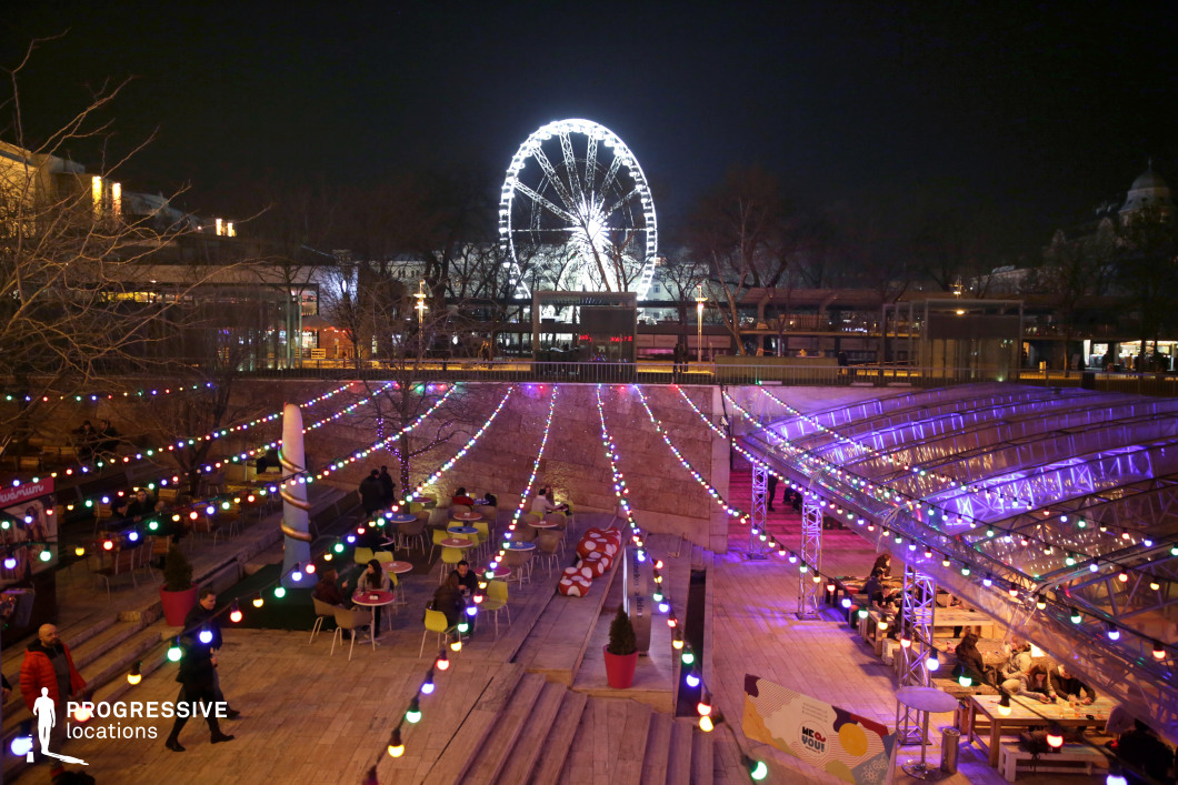 A lively urban nightlife scene unfolds in an open-air bar, illuminated by cascading strings of colorful lights. The multi-level seating area features modern, playful furniture, inviting guests to enjoy the dynamic ambiance. Overhead, a towering Ferris wheel glows brilliantly against the night sky, creating a striking visual focal point. The glass canopy on one side adds a contemporary architectural element, reflecting the vibrant hues of the lighting. The atmosphere is energetic yet intimate, making this location ideal for film scenes requiring a trendy social setting, festive gatherings, or romantic city nightlife sequences.