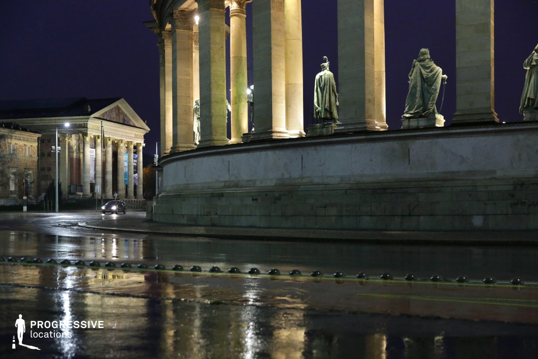 A grand neoclassical colonnade with towering stone columns and bronze statues dominates the scene, illuminated against the night sky. The statues, positioned on pedestals, depict historical figures draped in detailed, flowing garments, adding a regal and timeless quality to the composition. The colonnade’s structured symmetry and warm artificial lighting create a dramatic contrast with the dark blue evening sky. In the background, a classical-style building with an ornate pediment and large columns is visible, its facade adorned with banners suggesting a cultural or historical institution. The pavement in the foreground reflects the light, introducing an additional layer of depth and visual texture, emphasizing the grandeur of the setting. This location offers a visually rich and historically significant backdrop, ideal for period pieces, architectural photography, or cinematic sequences requiring a stately and dignified atmosphere. The interplay of light, shadow, and reflections enhances the monumental scale, making it a compelling choice for both historical and contemporary creative projects.