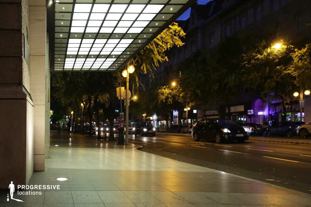 A sleek, contemporary hotel entrance with an illuminated canopy extends over a polished sidewalk, casting a soft, diffused glow onto the pavement. The geometric grid of lights in the canopy adds a modern and sophisticated architectural element, making the entrance visually prominent. The sidewalk is spacious, featuring embedded floor lights that subtly guide pedestrians along the building’s façade. Beyond the entrance, a lively urban street lined with trees and classic buildings stretches into the distance. The warm glow of streetlights reflects off the road, enhancing the dynamic atmosphere of the scene. Various illuminated storefronts, signage, and car headlights contribute to the bustling nighttime ambiance, evoking a vibrant city setting. This location is ideal for capturing urban nightlife, high-end hospitality settings, or contemporary city scenes. The contrast between the sleek, modern entrance and the historic architecture in the background makes it a versatile setting for film and photography, balancing luxury with an active metropolitan feel.