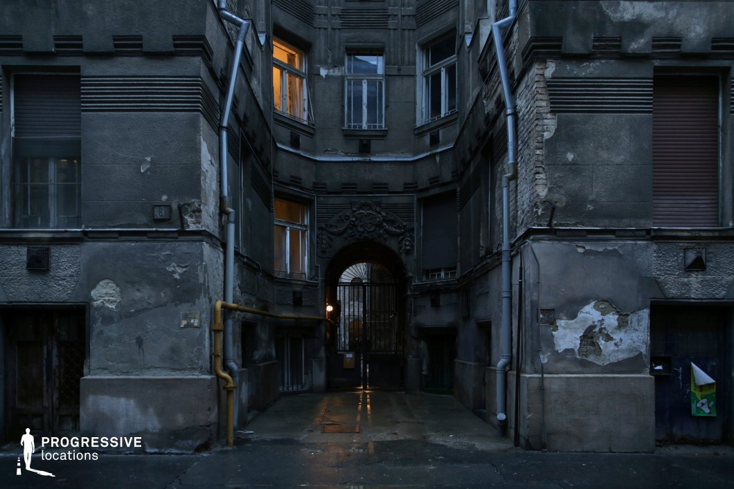 A weathered residential courtyard framed by deteriorating, early 20th-century architecture. The dark, textured facade exhibits peeling plaster, exposed brick, and visible signs of age, creating a raw, gritty atmosphere. Tall, narrow windows—some illuminated—break the monotony of the darkened exterior, hinting at life within. The heavy, wrought-iron gate at the arched passageway, adorned with intricate floral motifs, adds a touch of faded elegance to the otherwise stark environment. Metal pipes and gas lines run along the walls, their industrial presence contrasting with the structure's ornate yet eroded design. The pavement reflects a faint glow from a single light source, subtly emphasizing the depth of the passageway. The setting’s confined, almost claustrophobic composition makes it ideal for suspenseful or dramatic storytelling, offering a strong visual contrast between decay and lingering grandeur.