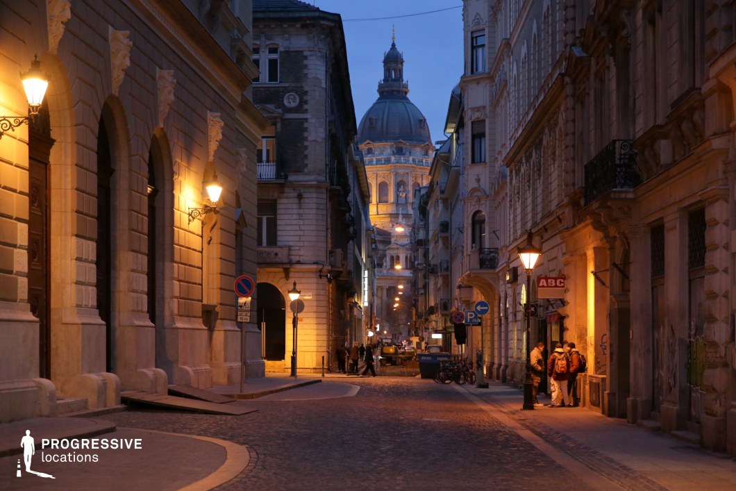 A picturesque cobblestone street winds through an elegant historic district, framed by grand neoclassical and baroque-style buildings. Ornate facades, intricate balconies, and sculpted details adorn the structures, exuding a timeless European charm. The warm glow of street lamps casts soft illumination on the stone facades, creating a rich interplay of light and shadow that enhances the architectural depth. In the background, a majestic domed basilica rises, bathed in golden light, serving as a dramatic focal point. The perspective of the narrow street naturally guides the eye toward this striking landmark, reinforcing the scene’s cinematic composition. The smooth curve of the cobblestone path and the symmetry of the buildings add to the harmonious aesthetic, making this location ideal for film productions, period dramas, or luxury brand visuals. This setting captures the essence of classic European cityscapes, blending historical grandeur with a welcoming, atmospheric warmth. Its rich architectural textures and refined ambiance make it perfect for sophisticated urban storytelling.