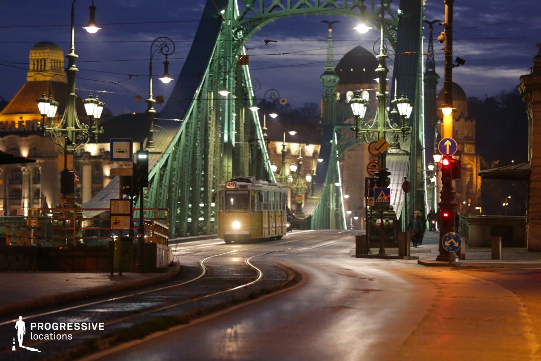 A striking green steel bridge with intricate architectural details spans across the cityscape, illuminated by a series of ornate street lamps that enhance its historic charm. The arched framework and decorative spires contribute to the bridge’s grandeur, while the tram lines running along its surface add an element of urban dynamism. A classic yellow tram approaches, its headlights cutting through the evening ambiance, reinforcing the scene’s timeless metropolitan character. In the background, historic domed buildings with illuminated facades complement the bridge’s ornate aesthetic, creating a layered composition rich in architectural heritage. The curved tram tracks and smooth road surface lead the eye naturally through the image, highlighting the bridge as a focal point. This location is ideal for urban cinematography, capturing the blend of historic and functional infrastructure. The strong geometric lines of the bridge, the atmospheric lighting, and the presence of public transportation make it a versatile backdrop for storytelling, fashion photography, or architectural studies.