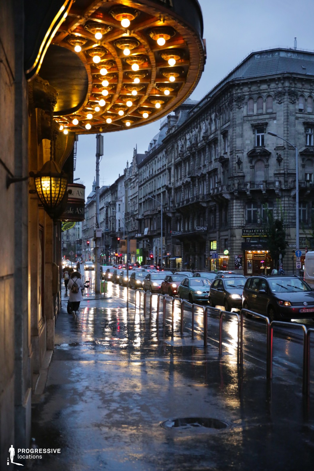 A grand theater or hotel entrance with an opulent, curved marquee extends over the sidewalk, adorned with rows of glowing light bulbs. The golden illumination contrasts with the cool tones of the historic city street, drawing attention to the building’s elegant architectural details. The vintage-style lanterns and decorative elements add a sense of old-world charm, making the entrance visually striking. Across the street, ornate stone buildings with intricate facades line the busy urban avenue, their presence reinforcing the city’s rich architectural heritage. The curved perspective of the street, along with the rows of traffic and shopfronts, creates a dynamic, cinematic depth. This location is ideal for capturing classic urban nightlife, historic luxury settings, or the atmospheric elegance of a cityscape that blends nostalgia with modern activity.