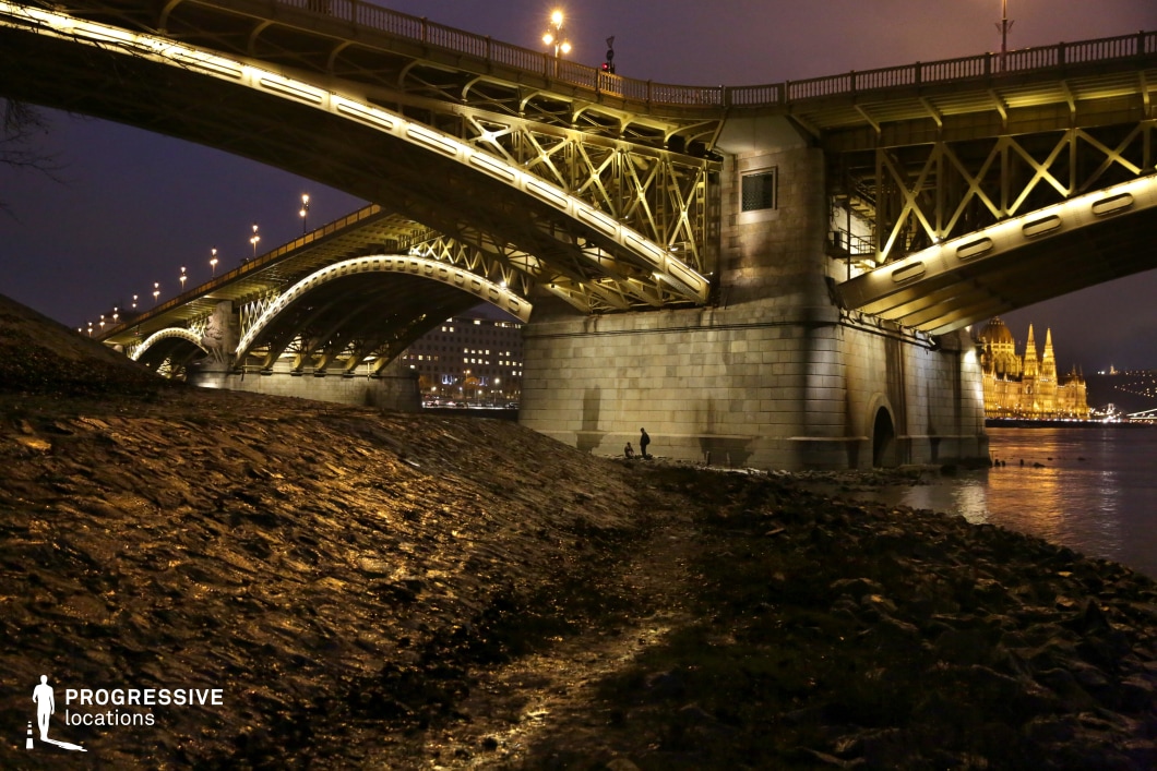A grand steel bridge with intricate metalwork and sturdy stone pillars stretches across the river, illuminated by a warm golden glow. The sweeping arches and crisscrossing support beams create a visually rich composition, drawing attention to the engineering marvel of the structure. The bridge's lighting highlights its detailed framework, emphasizing its elegance and historical significance. Below the bridge, a rocky riverbank adds texture to the foreground, leading the viewer's eye toward the water’s edge. In the distance, a majestic, illuminated neo-Gothic building with soaring spires dominates the skyline, enhancing the scene’s architectural depth and grandeur. The contrast between the monumental bridge and the serene riverside setting makes this location ideal for urban night photography, architectural studies, or cinematic sequences requiring a mix of structural brilliance and historic ambiance.
