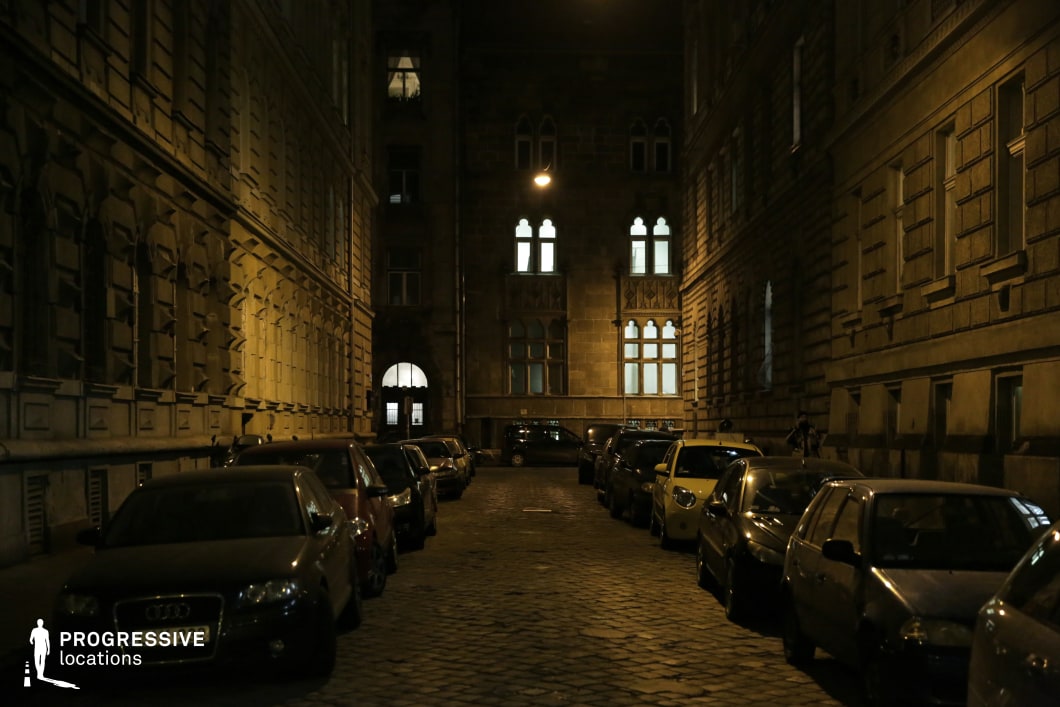 A dimly lit, narrow cobblestone street is enclosed by imposing historic buildings with intricate facades, decorative stonework, and tall arched windows. The soft glow of scattered streetlights highlights the texture of the aged masonry, casting long shadows that enhance the moody, atmospheric feel of the scene. The symmetry of the flanking structures guides the eye toward the illuminated entrance of a distant building, framed by a series of gothic-style windows that add an element of architectural elegance. The contrast between the warm artificial lighting and the dark, shadowed recesses of the buildings creates a visually dramatic composition. The combination of old-world charm and subdued lighting makes this location well-suited for noir-style cinematography, urban photography, or narratives that require a secluded yet richly detailed European street setting.