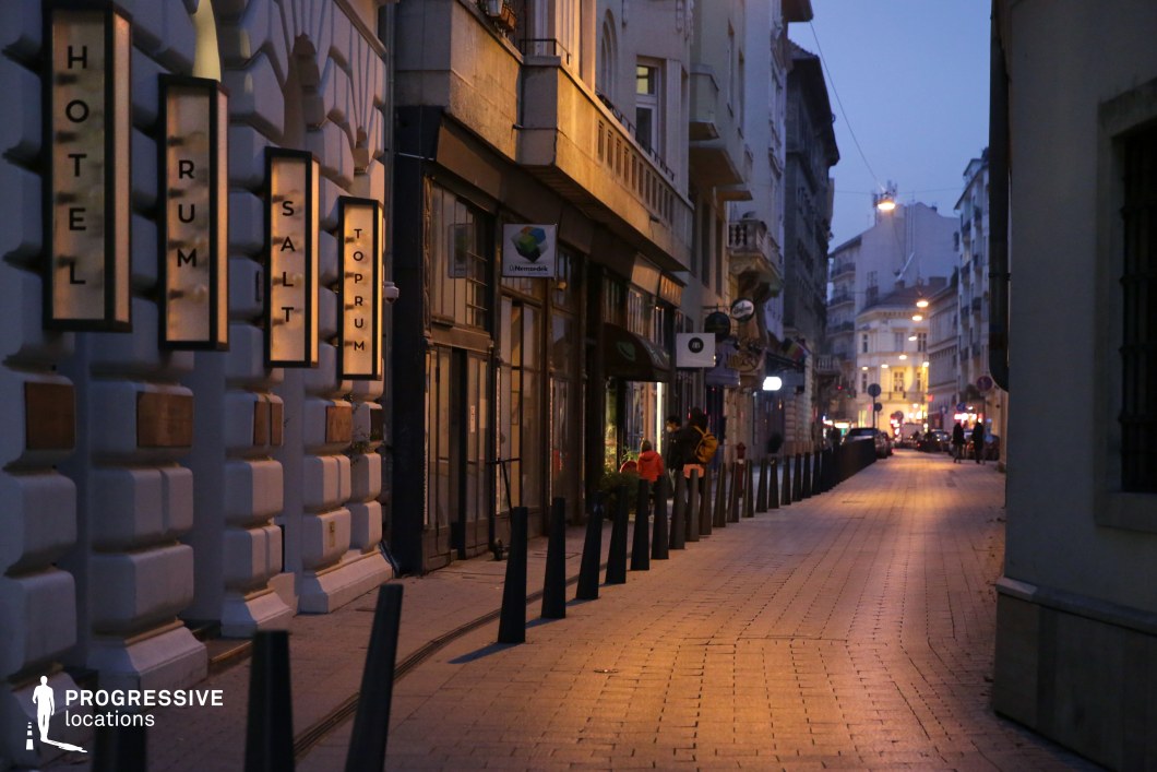 A picturesque, pedestrian-friendly side street lined with elegant historic buildings and boutique storefronts. The softly illuminated facades display intricate architectural details, including decorative moldings, balconies, and large windows that add depth and character to the scene. A row of evenly spaced bollards guides the eye along the gently curving cobbled path, enhancing the street’s structured yet inviting feel. On the left, a refined hotel entrance features vertically arranged illuminated signage, contributing to the warm ambiance. The street is active yet intimate, with distant figures and shop windows glowing, creating a sense of urban vibrancy. The background reveals a brightened intersection, where the street opens into a larger square, adding a sense of depth and movement to the composition. This location is ideal for sophisticated city visuals, lifestyle photography, or cinematic sequences that require a refined yet lively European streetscape. Its blend of historical charm and modern boutique culture makes it a versatile and visually compelling urban setting.