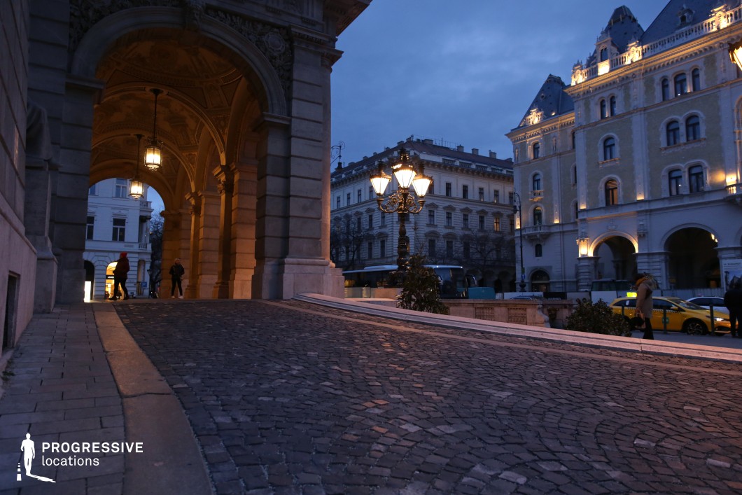 A stately arched passageway with intricate classical detailing frames this cobblestone path, leading into a refined European square. The vaulted ceiling of the arch is adorned with ornate carvings, while hanging lanterns provide a warm, atmospheric glow that contrasts with the cool twilight sky. The stone facade exudes grandeur, reinforcing the architectural richness of the setting. Beyond the arch, an elegant square lined with historic buildings unfolds. The softly illuminated facades feature arched walkways, decorative cornices, and steeply pitched roofs, showcasing a blend of classical and neo-Gothic influences. A vintage-style street lamp in the foreground adds a timeless charm, further enhancing the sophisticated ambiance of the scene. This location is ideal for period dramas, historical storytelling, or luxury brand visuals. The depth created by the archway, combined with the upscale architectural elements, makes it a visually compelling backdrop for cinematic compositions.