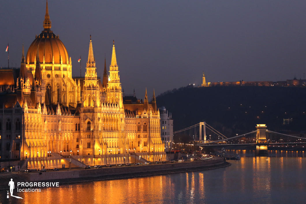 A breathtaking neo-Gothic architectural masterpiece dominates the riverbank, its intricate spires and grand central dome glowing warmly against the evening sky. The elaborate detailing of the facade is accentuated by the soft golden lighting, emphasizing the structure’s historical significance and regal presence. The shimmering reflections on the river enhance the visual depth, making the scene feel both majestic and serene. Beyond the landmark, a suspension bridge adorned with elegant lights spans the river, seamlessly connecting different parts of the city. In the background, a hilltop monument stands illuminated, adding another layer of grandeur to the composition. The careful balance of natural and artificial light creates a cinematic atmosphere, making this location ideal for capturing historic elegance, architectural grandeur, or the refined beauty of a European riverside cityscape.