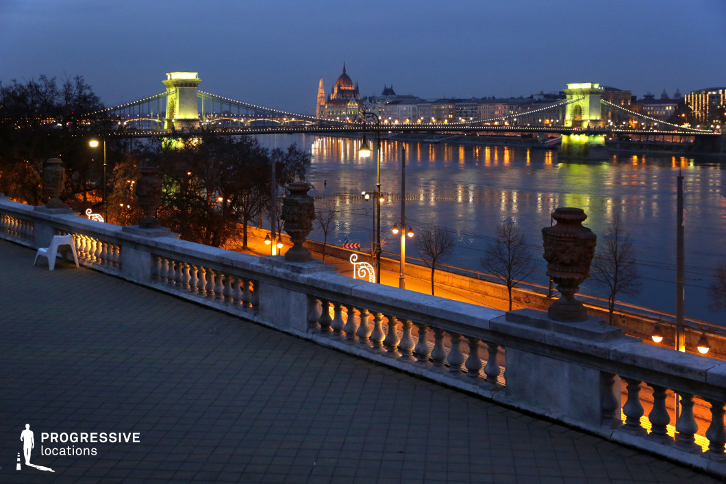 An elevated terrace with classical stone balustrades overlooks a stunning riverside panorama, featuring a gracefully illuminated suspension bridge stretching across the water. The bridge’s elegant arches and glowing lights create a captivating visual rhythm, while the reflections on the river enhance the sense of depth and grandeur. Ornate decorative urns along the terrace add an element of historic sophistication, reinforcing the refined character of the location. Beyond the bridge, a magnificent neo-Gothic building rises prominently against the evening sky, its intricate facade bathed in warm golden light. The softly glowing street lamps along the promenade below cast a welcoming ambiance, guiding the eye through the scene’s rich architectural layers. The combination of the elevated vantage point, iconic landmarks, and atmospheric lighting makes this setting ideal for luxury visual compositions, cinematic cityscape captures, or refined urban storytelling.