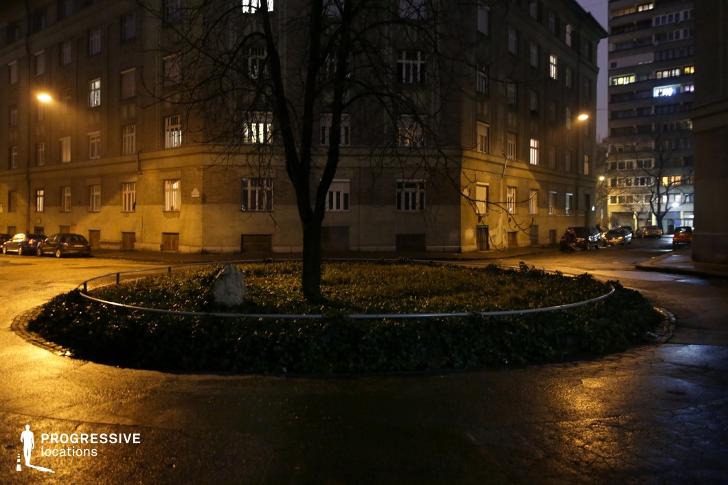 A modest, circular green space with a single tree and low vegetation sits at the center of a quiet residential roundabout. The surrounding buildings, with their uniform facades and simple architecture, evoke a classic Eastern European urban atmosphere. The soft, yellow glow of streetlights casts long shadows, accentuating the subdued and structured nature of the scene. In the background, a mix of mid-century and modern apartment buildings adds depth to the urban setting. The curved road encircling the roundabout creates a natural focal point, leading the viewer’s eye through the space. This location offers a versatile setting for understated urban storytelling, noir-style cinematography, or scenes requiring an unembellished yet evocative cityscape.
