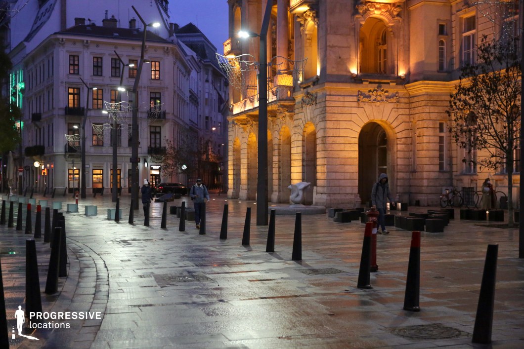 A sophisticated urban square framed by grand neoclassical buildings, featuring ornate facades, arched entrances, and decorative stone balconies. The golden illumination of the historic structure contrasts beautifully with the cool blue hues of the evening sky, highlighting intricate architectural details. The presence of scattered black bollards and modern street lighting blends historic grandeur with contemporary urban design. The smooth, tiled pavement extends into the distance, bordered by stylish storefronts and classic European buildings with large windows and elegant balconies. The carefully arranged urban elements, including trees and sculptural accents, enhance the refined and balanced aesthetic of the space. This location offers a versatile setting for high-end urban cinematography, period productions, or luxury fashion photography. The blend of classical architecture and modern urban planning makes it ideal for capturing sophisticated European cityscapes.