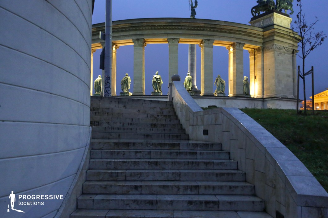 A striking neoclassical monument stands illuminated against the evening sky, framed by an elegant curved colonnade adorned with bronze statues of historical figures. The grand staircase in the foreground, made of smooth stone with a gracefully curving handrail, leads towards the towering structure, enhancing the sense of depth and perspective. The monument’s architectural symmetry and intricate sculptural details create a visually commanding presence. The contrast between the structured stonework and the surrounding greenery adds balance to the composition, while the soft artificial lighting emphasizes the monument’s grandeur. The positioning of the statues, each in dignified poses, reinforces the site's historical significance. This location is ideal for capturing monumental architecture, historical reenactments, or cinematic compositions requiring a regal and timeless urban backdrop. The interplay of light, texture, and perspective makes it a compelling setting for dramatic storytelling or refined visual narratives.