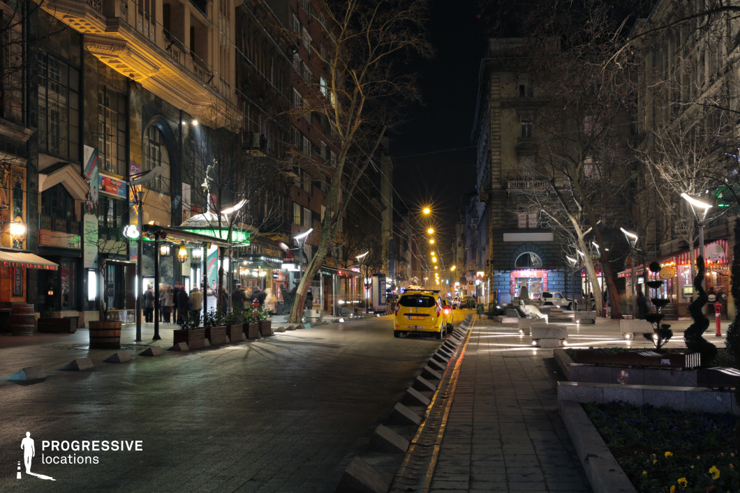 A lively, illuminated city street lined with a mix of historic and modern buildings, featuring elegant facades, large windows, and decorative balconies. The warm glow of streetlights stretches into the distance, creating a visually striking perspective that emphasizes the street’s depth and urban energy. The combination of modern LED fixtures and traditional lantern-style lighting enhances the dynamic contrast between old and new architectural styles. The street is active with storefronts, theaters, and cafés, adding a cultural and artistic flair to the scene. A yellow taxi stands prominently in the foreground, reinforcing the bustling, cosmopolitan atmosphere. Sculpted greenery and well-placed street furniture contribute to the stylish and pedestrian-friendly design of the area. This location is ideal for capturing the vibrancy of city nightlife, urban elegance, and cultural hotspots. Its rich architectural diversity, atmospheric lighting, and cinematic depth make it a perfect backdrop for high-end fashion photography, film productions, or sophisticated urban storytelling.