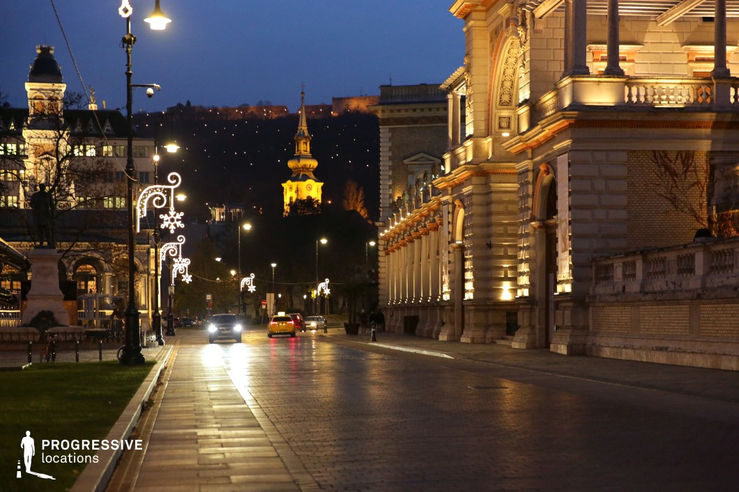 A grand European boulevard lined with historic buildings featuring intricate facades, arched entrances, and decorative columns, all bathed in soft golden lighting. The wide cobblestone-paved street leads towards an illuminated church tower in the background, which rises prominently against the dark hillside, creating a visually striking focal point. Elegant street lamps adorned with delicate ornamental details add a refined touch to the scene, complementing the classical architecture. The interplay of warm building lights and cooler ambient street lighting enhances the depth and contrast of the setting, making it an inviting yet sophisticated urban environment. This location is ideal for luxury urban photography, high-end brand visuals, or cinematic compositions requiring a blend of classical architecture and atmospheric city lighting. The rich textures, historical depth, and well-balanced lighting make it a versatile and visually engaging setting for refined storytelling.