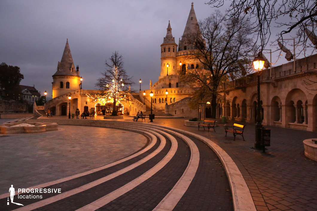 An enchanting medieval-style plaza unfolds with curved stone steps leading toward grand limestone towers adorned with arched windows and turrets. The warm golden lighting enhances the architectural elegance, casting a soft glow against the cloudy evening sky. Delicate arches and intricate stonework lend a fairy-tale quality to the setting, creating a balance between history and fantasy. A beautifully illuminated tree stands at the heart of the square, adding warmth and festive charm to the scene. Elegant wrought-iron street lamps line the pathways, complementing the historic ambiance. Wooden benches and neatly arranged cobblestone walkways enhance the inviting nature of the space, making it a perfect setting for quiet reflection or cinematic storytelling. This location is ideal for period dramas, fantasy narratives, or high-end travel photography. Its combination of architectural grandeur, atmospheric lighting, and historic charm makes it a visually stunning backdrop for sophisticated and timeless storytelling.