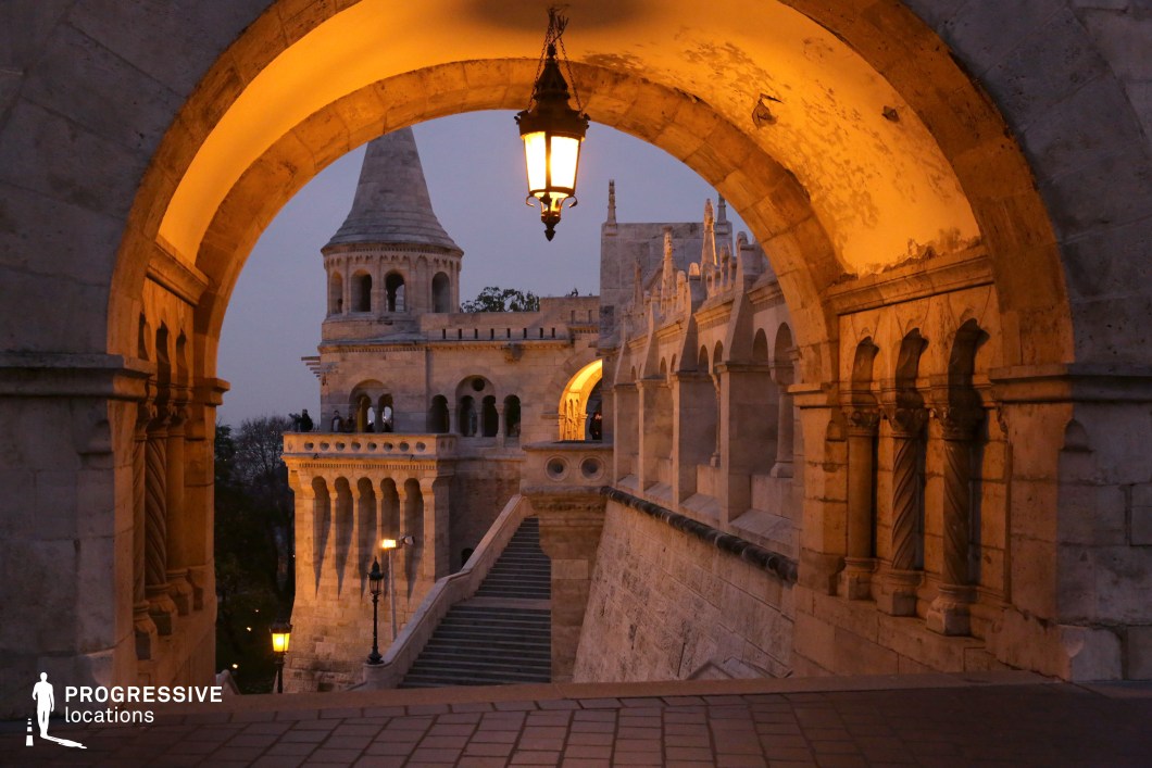 A grand medieval-style limestone tower is beautifully framed by an elegant stone archway, creating a striking sense of depth and perspective. The hanging lantern at the center emits a warm glow, contrasting with the cool evening sky and highlighting the intricate details of the carved stone. The scene evokes a timeless, almost fantastical ambiance, with the softly illuminated staircases and arched walkways adding to its cinematic quality. The sweeping stairway leads upward along a series of elegant colonnades, adorned with delicate carved patterns and sculpted details. The interplay of light and shadow enhances the dramatic curves of the architecture, making this location feel both intimate and monumental. This setting is ideal for historical films, fantasy storytelling, or luxury travel photography. The refined stone craftsmanship, balanced lighting, and immersive medieval charm make it a perfect backdrop for visually captivating narratives.