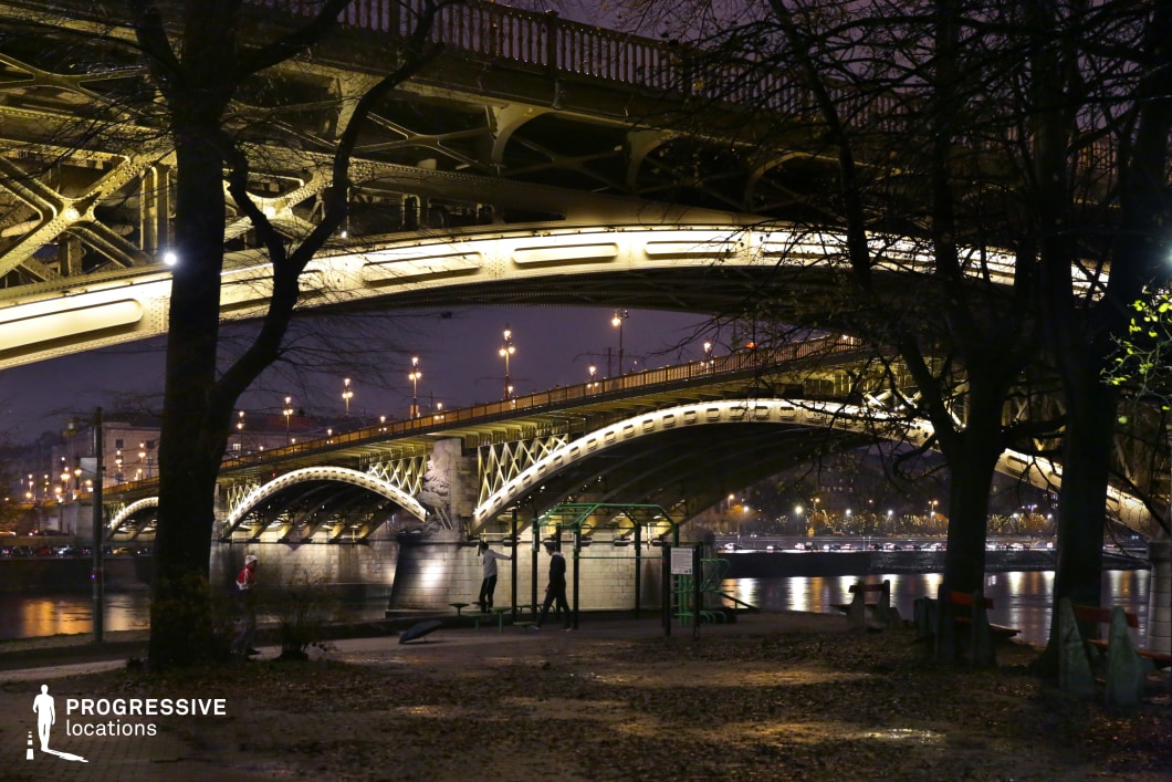 A grand steel bridge with intricate ironwork spans across the river, its elegant arches glowing under warm artificial lighting. The bridge’s layered structure, highlighted by a series of evenly spaced lights, creates a visually compelling geometric pattern. Beneath it, the river reflects the golden illumination, adding depth and movement to the composition. In the foreground, a small riverside park with leafless trees and scattered benches provides a tranquil contrast to the bridge’s grandeur. The soft interplay of shadows and artificial lights enhances the nighttime atmosphere, making the scene feel cinematic and immersive. This location is ideal for urban photography, architectural cinematography, or storytelling that captures the contrast between monumental engineering and intimate city life. The combination of industrial elegance and natural surroundings creates a versatile setting for visually striking compositions.