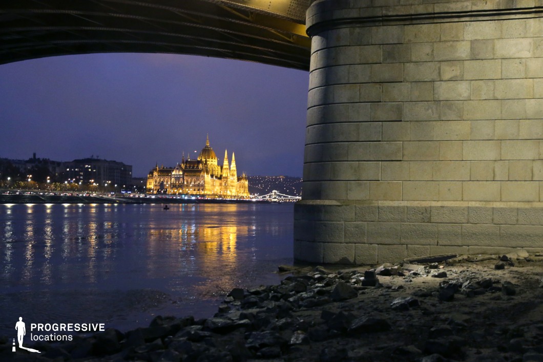 A grand architectural landmark glows in golden light, standing prominently along the riverbank, framed dramatically by the underside of a bridge. The massive stone pillar in the foreground adds depth and structure to the composition, contrasting with the shimmering reflections on the water. The bridge’s underside and its industrial framework create a dynamic visual frame that enhances the perspective toward the distant illuminated building. The riverside setting, with its rocky shoreline and textured stone surfaces, adds a rugged contrast to the refined architecture in the background. The balance of warm artificial lighting and cool twilight tones in the sky contributes to the cinematic quality of the scene. This location is ideal for atmospheric urban cinematography, architectural photography, or visually compelling compositions that contrast natural and man-made elements. The combination of structural framing, dramatic lighting, and reflections creates a striking and immersive setting.
