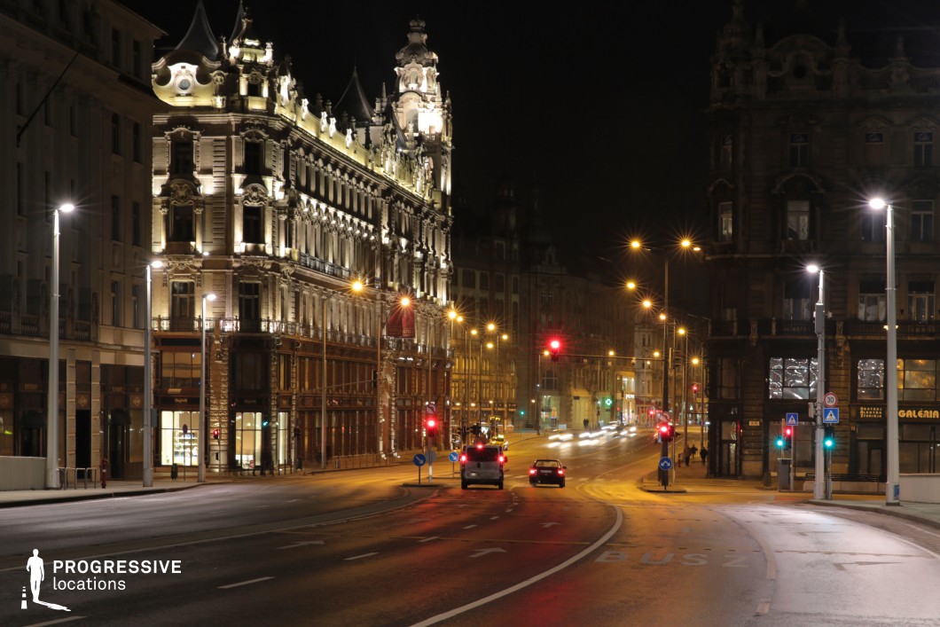 A grand historic boulevard stretches into the distance, lined with ornate, illuminated buildings that showcase intricate architectural details. The contrast between the glowing facades and the dark night sky enhances the elegance of the scene. The wide, curving street is lined with evenly spaced streetlights, creating a rhythmic visual pattern that guides the eye toward the horizon. The scene captures the interplay of old-world grandeur and modern infrastructure, with well-maintained roads, visible traffic markings, and a network of glowing traffic signals. The reflections of artificial lights on the smooth asphalt add depth and texture to the composition. This setting is ideal for film productions, nighttime urban photography, and atmospheric cityscape visuals. The combination of grand architecture, urban lighting, and a dynamic perspective creates a compelling and cinematic backdrop.