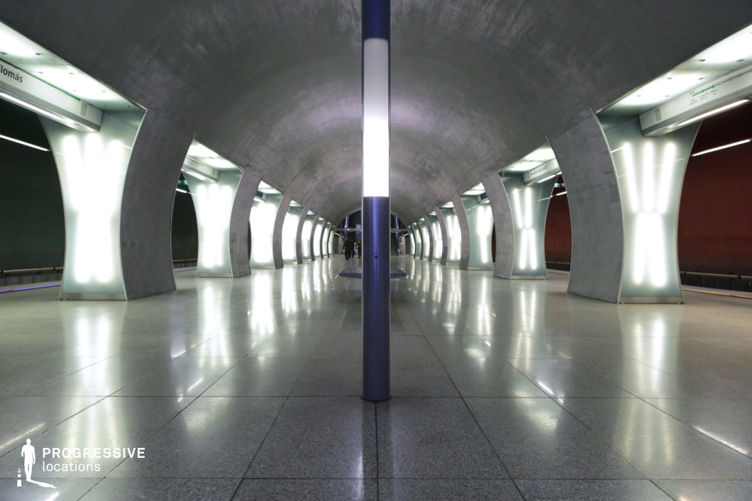 The image showcases a futuristic underground platform with a sleek and industrial design. The space is characterized by smooth, metallic, and concrete surfaces that reflect light, creating a sense of spaciousness. The prominent feature of this design is the row of luminous, curved pillars that run along the platform. These pillars, which serve both as structural elements and sources of light, are encased in translucent material, giving them an ethereal glow. The floor is polished and smooth, adding to the modern, high-tech feel of the space. The overall atmosphere is clean, minimalist, and open, with a sense of symmetry and linearity that guides the viewer's eye down the platform. This location is well-suited for urban or industrial scenes, offering a visually striking environment with a contemporary architectural style.
