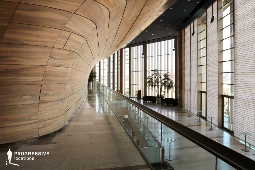 The image captures the striking interior of a contemporary architectural space, defined by sweeping curves and the natural elegance of wood. A smoothly curved wooden wall dominates the scene, creating a dynamic flow that contrasts with the sleek glass railing running along the corridor. The wood panels are arranged in a visually appealing pattern, enhancing the natural warmth and texture of the material. On the left side, tall windows allow natural light to flood the space, offering a panoramic view of the exterior. Potted plants stand gracefully by the windows, adding a touch of greenery to the minimalist design. The spacious, light-filled environment is balanced by the intricate details of the architecture, which include large glass walls adorned with subtle script-like patterns, contributing to the space's sophisticated and artistic ambiance. This setting is ideal for high-end events, modern art exhibitions, or elegant social gatherings, combining functionality with aesthetic appeal in an urban environment.