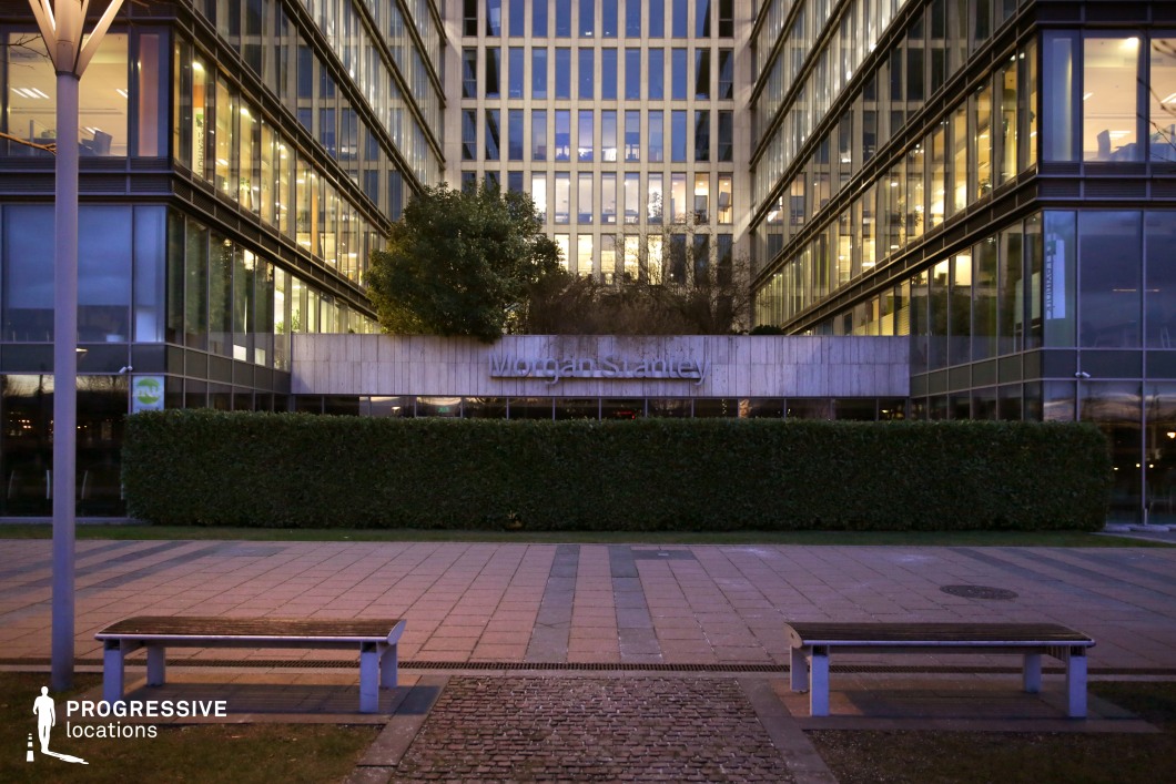 The image showcases the sleek exterior of a modern office building, characterized by its clean lines and geometric design. The building’s facade is primarily made of glass, with large, reflective windows that create a sense of openness and transparency. A prominent concrete wall at the base features the 