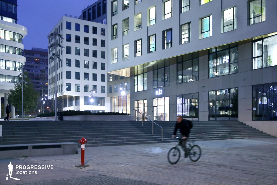 The image captures a modern urban plaza in the evening, where clean lines and functional design meet. Two contemporary office buildings stand side by side, featuring large, square windows that illuminate the space, creating a vibrant atmosphere. The structures are minimalistic, with white facades and simple architectural details. In the foreground, a wide staircase leads up to the entrance, with its smooth steps offering an accessible route into the building. On the right side, a small shop with illuminated signage adds life to the area, contributing to the city’s nighttime energy. A cyclist passes by, adding a sense of motion to the tranquil scene. A red fire hydrant and strategically placed street lamps enhance the urban aesthetic, casting soft lighting on the surrounding pavement. The juxtaposition of geometric architectural elements with the open space creates a balanced and welcoming setting for various activities, making it suitable for modern office environments or urban gathering spaces. The lighting and modern structures evoke a sense of cleanliness and order, ideal for commercial and professional settings.