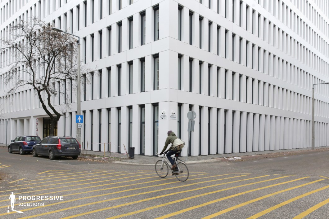 This image features a modern office building with a minimalist design, highlighted by its clean, white facade and orderly, grid-like window arrangement. The building’s sharp, geometric edges emphasize its sleek, contemporary style. The structure is located on a city corner, where the building’s lines form a distinct intersection. On the street level, a cyclist rides across a wide, yellow-striped crosswalk, adding a sense of movement to the scene. Nearby, two cars are parked along the curb, while a single tree provides a natural contrast to the surrounding architecture. The overall atmosphere reflects a quiet, urban environment, with an emphasis on functionality and order. The design of the building and surrounding streets makes it an ideal location for business or professional settings, with its clean and straightforward aesthetic supporting a variety of uses.
